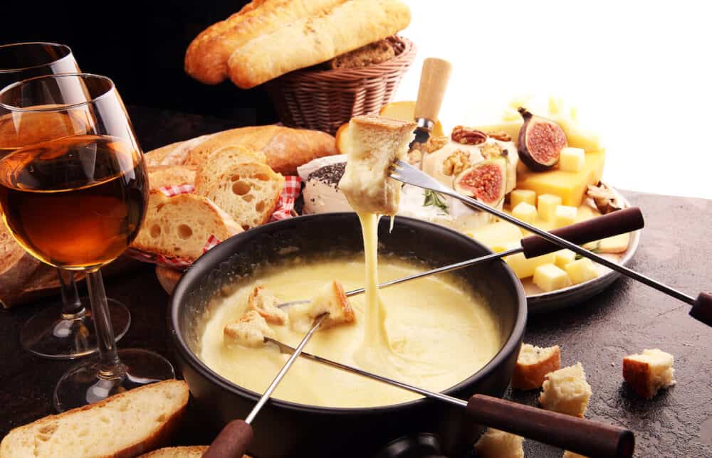 Gourmet Swiss fondue dinner on a winter evening with assorted cheeses on a board alongside a heated pot of cheese fondue with two forks dipping bread