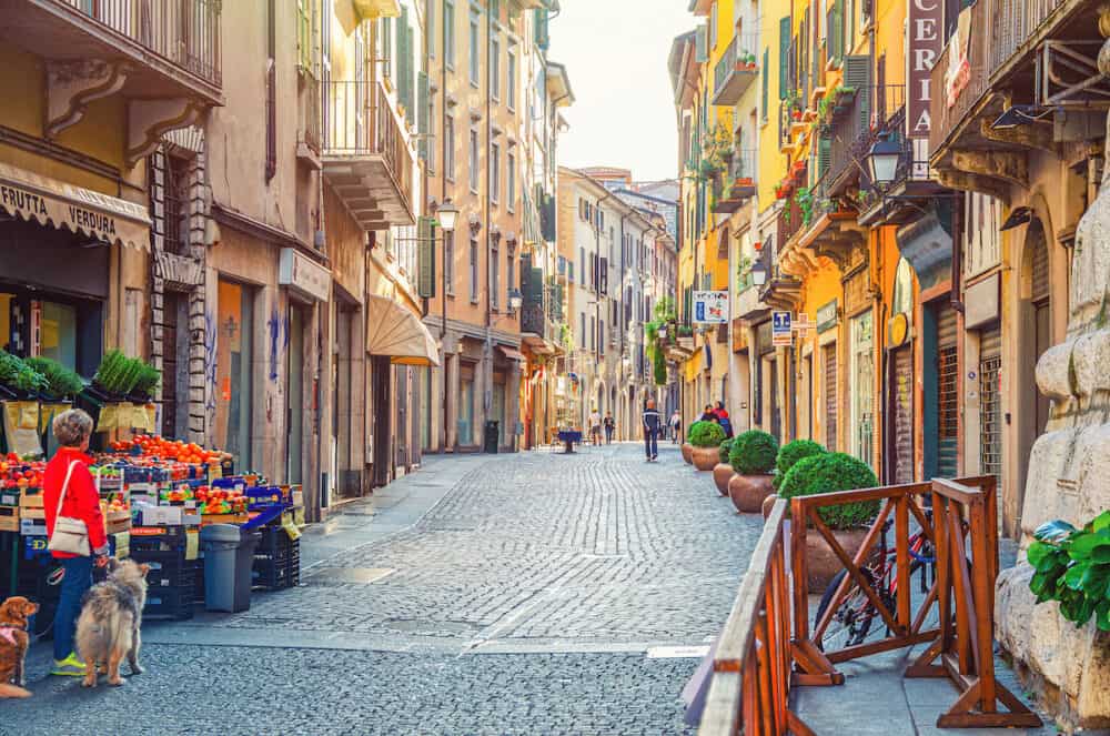 Brescia, Italy -  Typical italian narrow street with traditional old buildings, cobblestone road, fruit and vegetables store and walking people, historical city centre, Lombardy