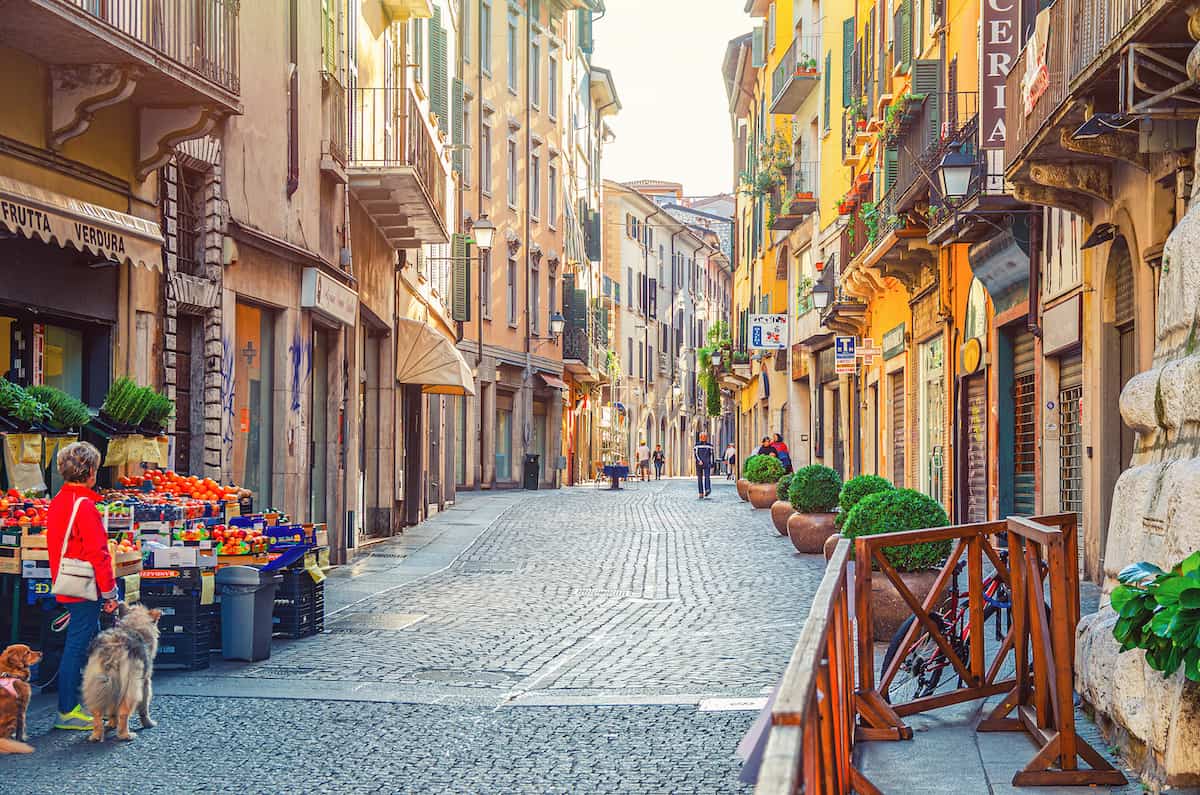 Brescia, Italy - Typical italian narrow street with traditional old buildings, cobblestone road, fruit and vegetables store and walking people, historical city centre, Lombardy
