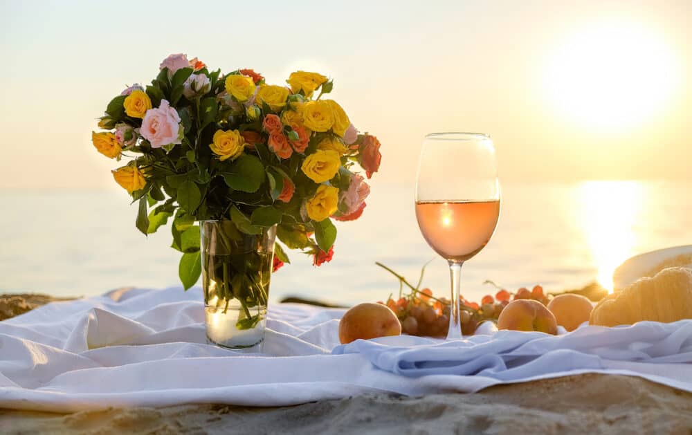 Romantic picnic at sunset by the sea on the beach, focus on glass. Delicate bouquet of flowers and sweet snacks