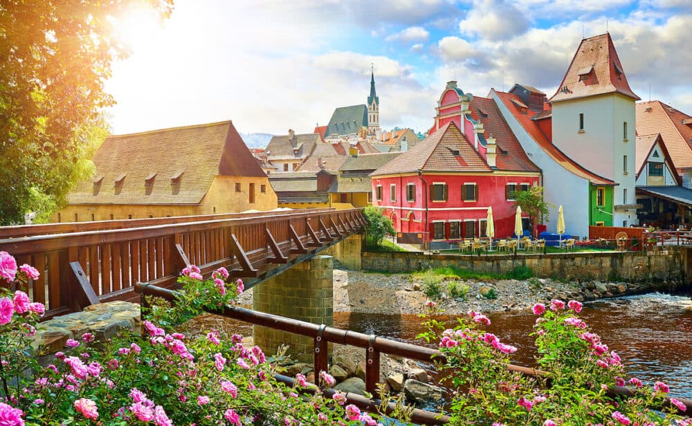 Czech Krumlov, (Cesky Krumlov), Czech Republic. Wooden bridge over river Vltava. Vintage picturesque old town with colorful houses and chapel of church. Rose flowers on bank. Sunny summer day.