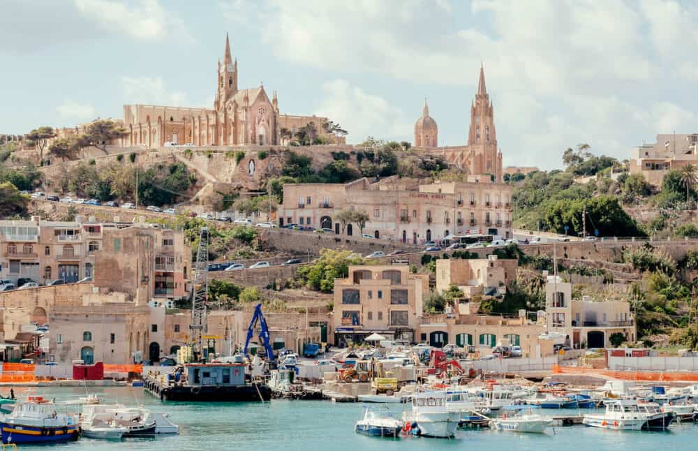 GOZO, MALTA: Gozo island port view with fishing boats and old churches on a hill. Mediterranean Sea island Gozo has population near 35,000