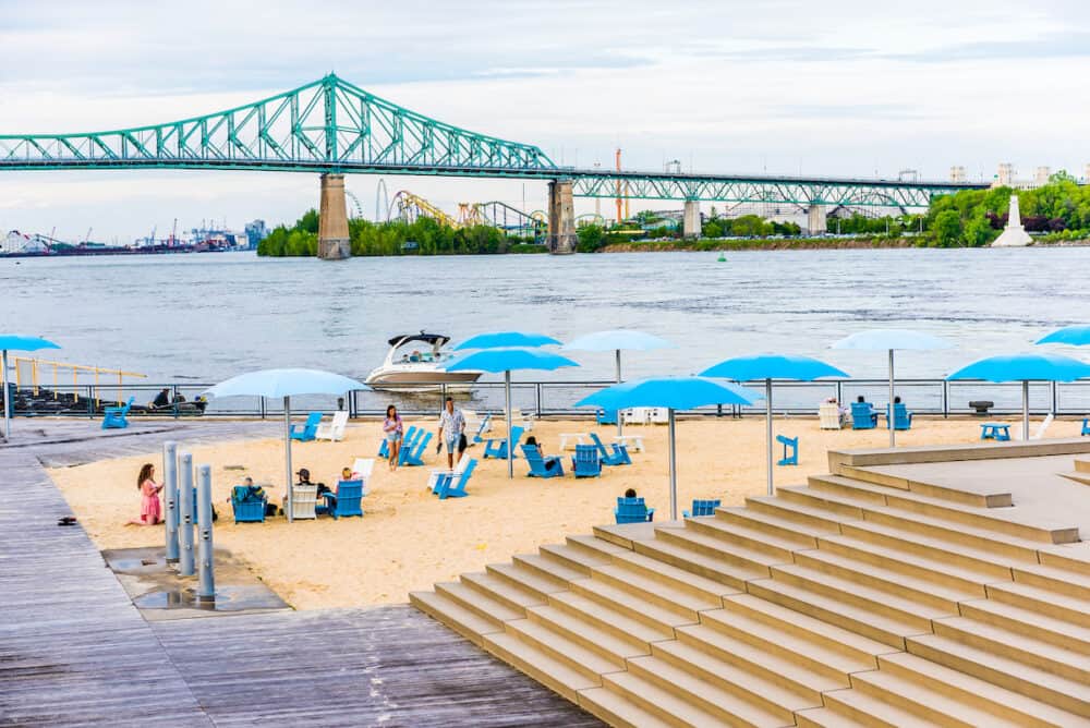 Old port area with sandy beach umbrellas and boats in city in Quebec region during sunset with Jacques Cartier bridge