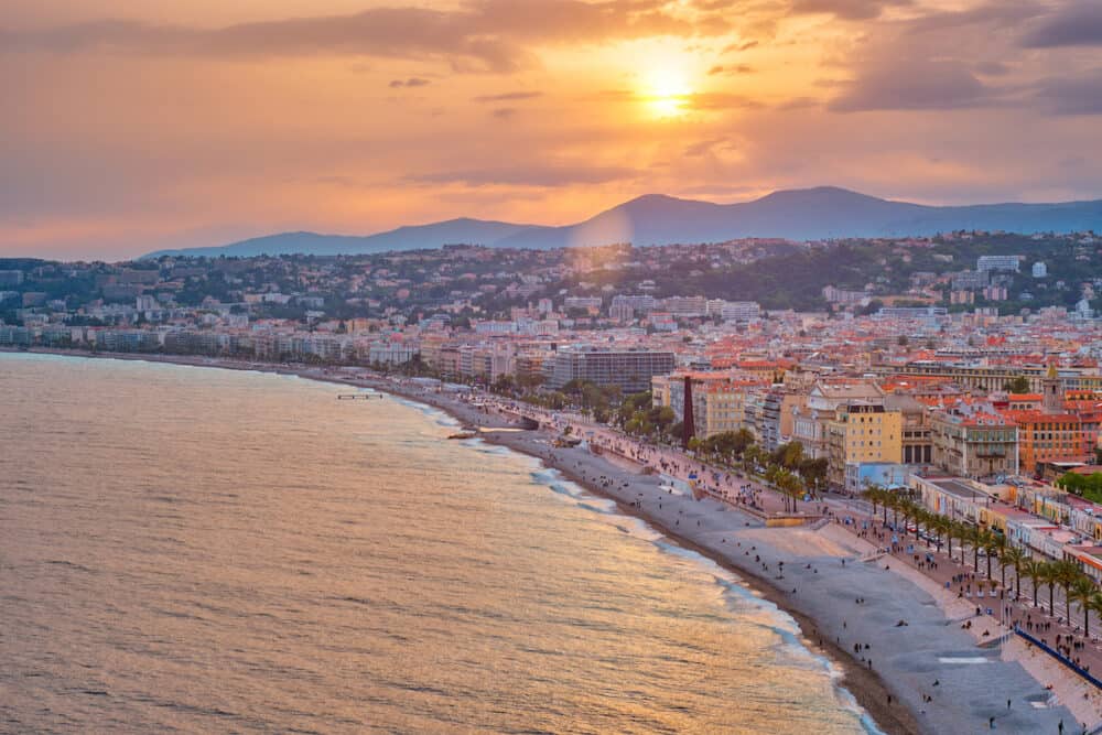 Picturesque scenic view of Nice, France on sunset. Mediterranean Sea waves surging on the beach, people are relaxing on the beach, cars driving the road. Nice, France