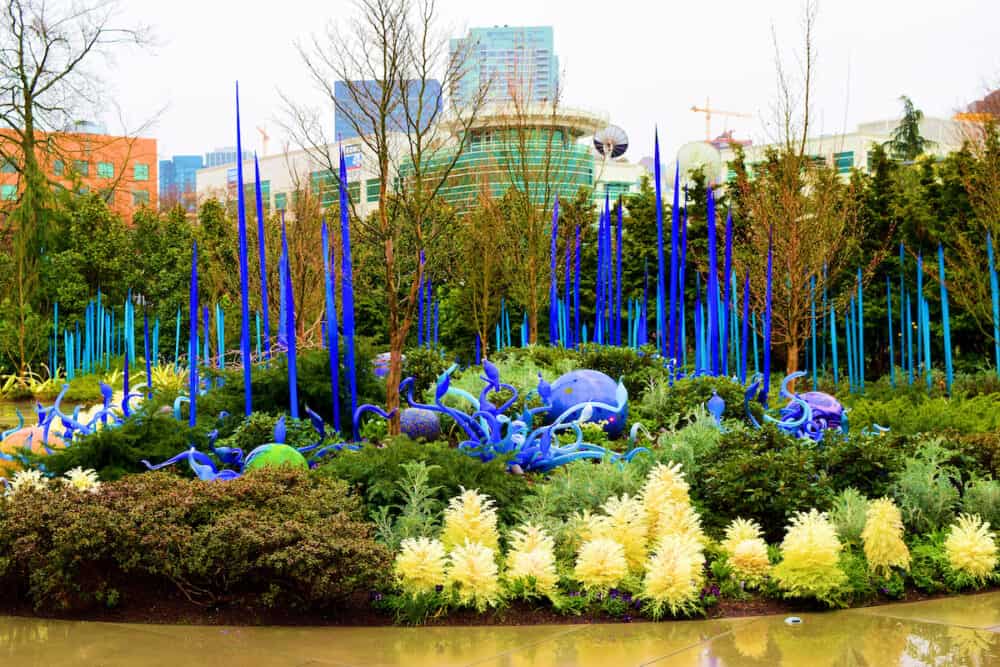 Seattle, WA:  Plants and flowers amongst glass art sculptures with the Seattle Skyline beyond taken at the Chihuly Garden and Glass Museum in Downtown Seattle, WA where people can see manicured gardens surrounded by glass art sculptures