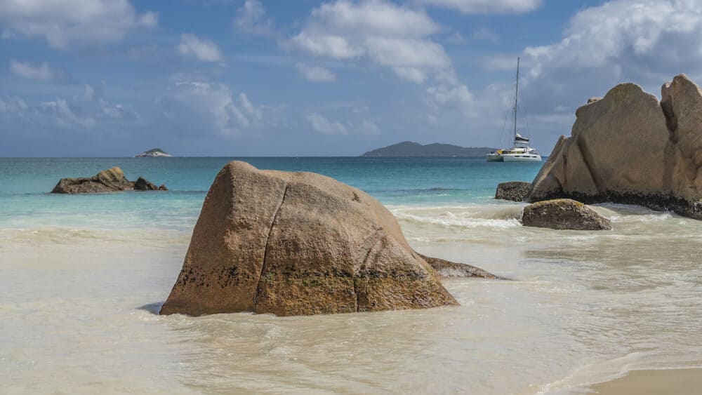 Picturesque granite rocks in the surf. The waves of the turquoise ocean spread over the sandy beach. The yacht and the silhouette of the island in the distance. Clouds in the blue sky. Seychelles