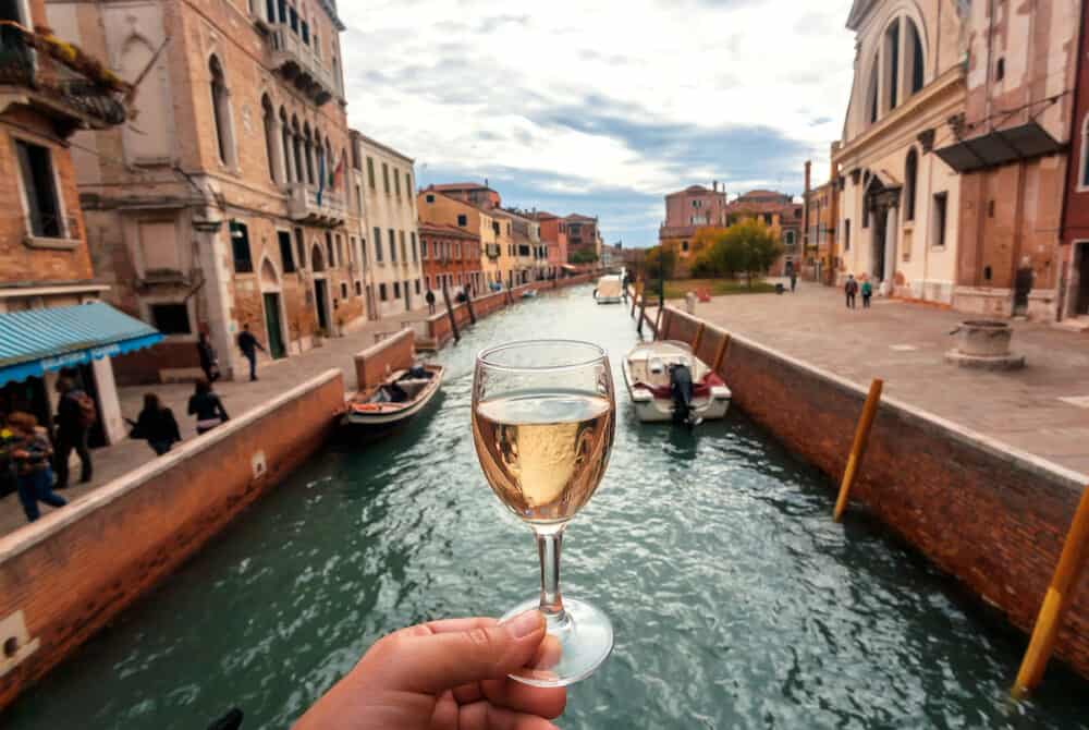 VENICE, ITALY: Water canals in Venice and white wine glass in hand of happy tourist on 8 October 2021. Ancient italian city and lagoon are UNESCO World Heritage Sites