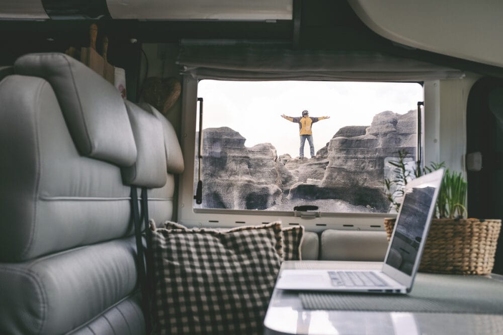 Digital nomad travel people lifestyle and alternative office with outdoors freedom. Laptop inside camper van. Man standing happy and free outside on a rock. Remote worker enjoying happiness outside
