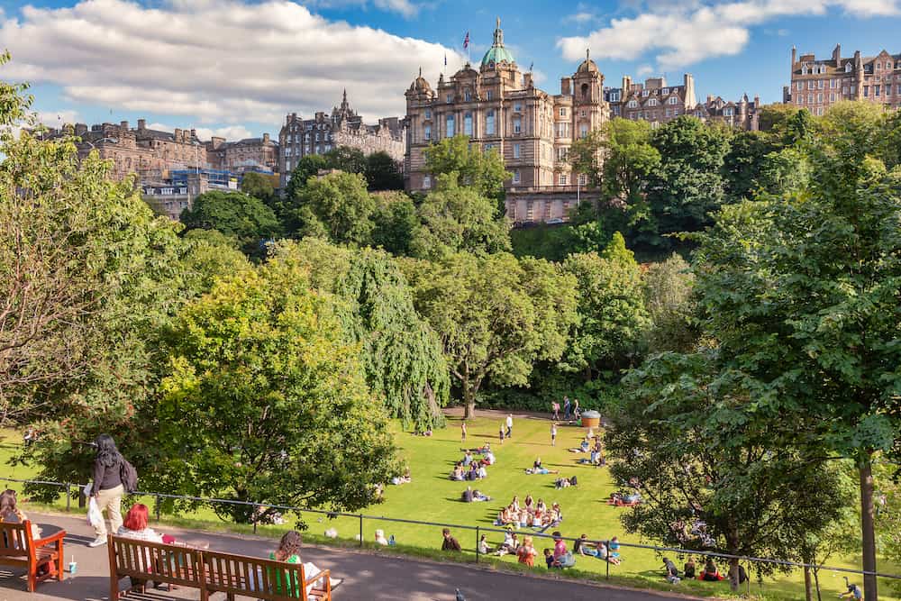 Locals and tourists enjoy warm sunny summer day relaxing and picnicking at the Princes Street Gardens, a famous public park in the city center