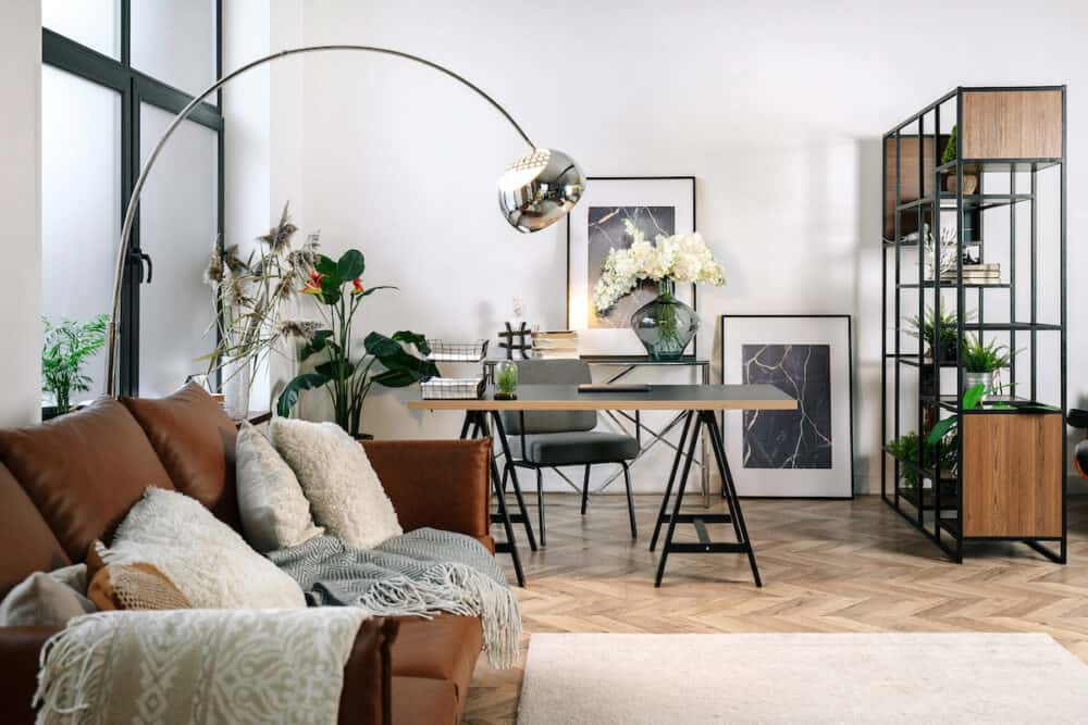 Home office in living room with modern interior design, comfort sofa, laptop computer, paper documents on work desk near armchair. Cozy apartment with decor, picture, bouquet in vase and houseplants