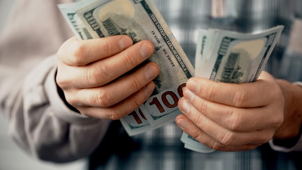Hands of man in blue t-shirt counting US Dollar bills or paying in cash on money background. Concept of investment, success, financial prospects or career advancement