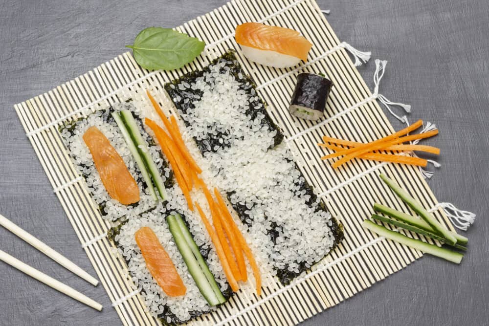 Process of making sushi. Rice, chopped carrots and cucumbers, salmon slices on bamboo mat. Flat lay. Grey background.