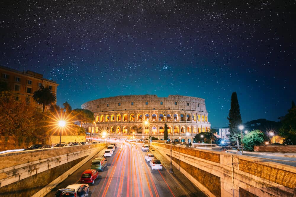 Rome, Italy. Colosseum Also Known As Flavian Amphitheatre In Evening Or Night Time. Night Traffic Light Trails Near Famous World Landmark. Bright Blue Night Sky. Amazing Night Starry Sky Background.