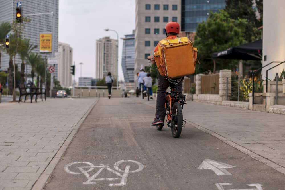 A pizza delivery man with a yellow backpack and in a red helmet rides a bicycle on a bicycle path with white markings on the main street of the city