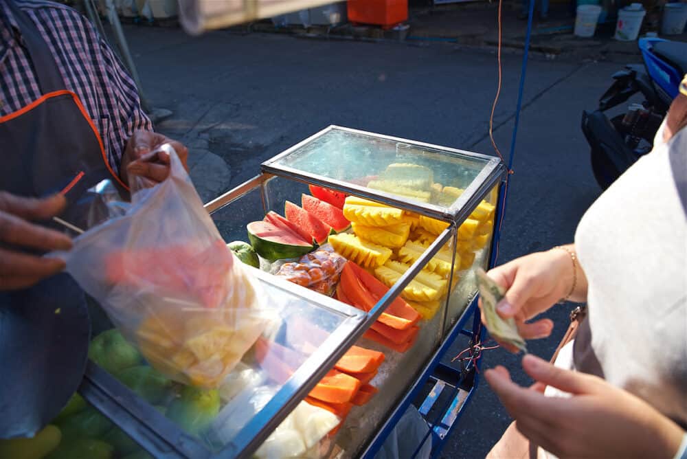 tropical fruit street vendor on the streets of Bangkok in Thailand, slicing different fruits, a woman gives money to a seller, selling from a tray