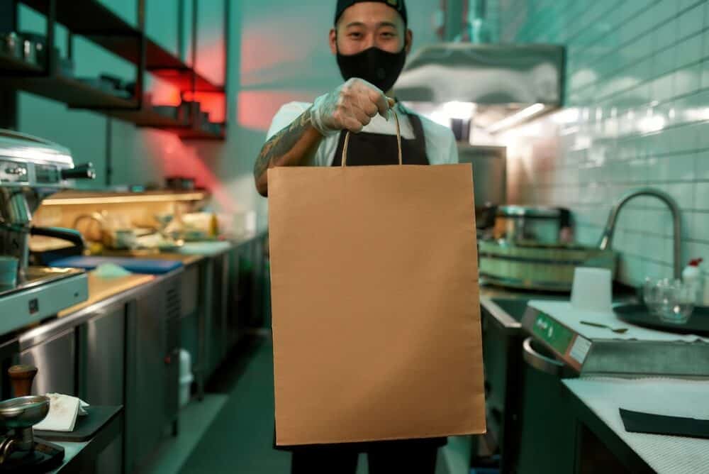 Young man standing in a restaurant with a large package of food Food for delivery during quarantine days Masked man in the restaurant kitchen with a package of food