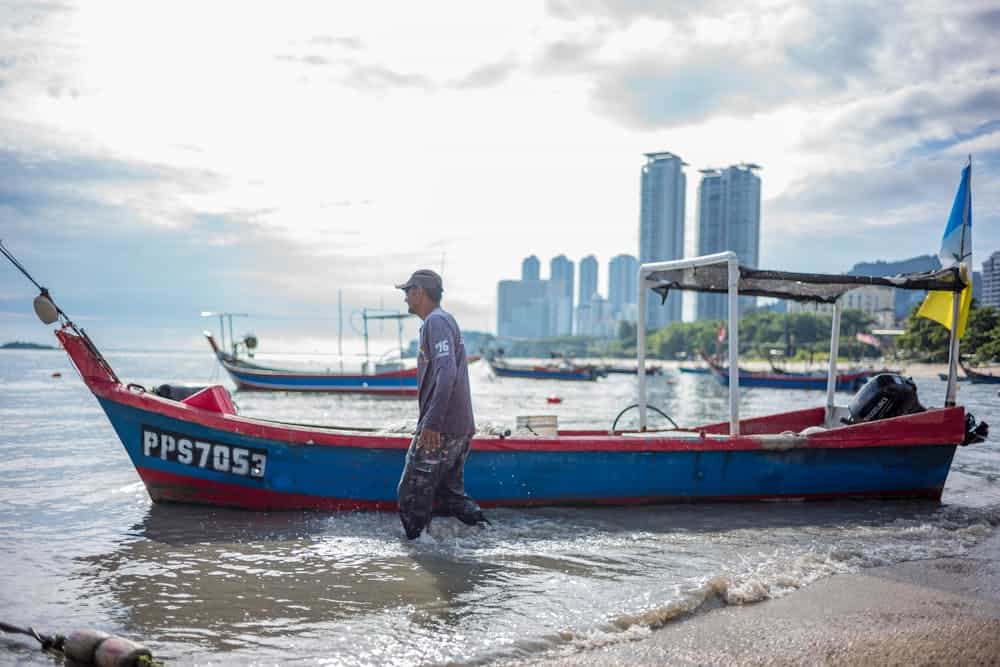 Fisherman walks near a boat on the shore with a city skyline backdrop in George Town, Malaysia.